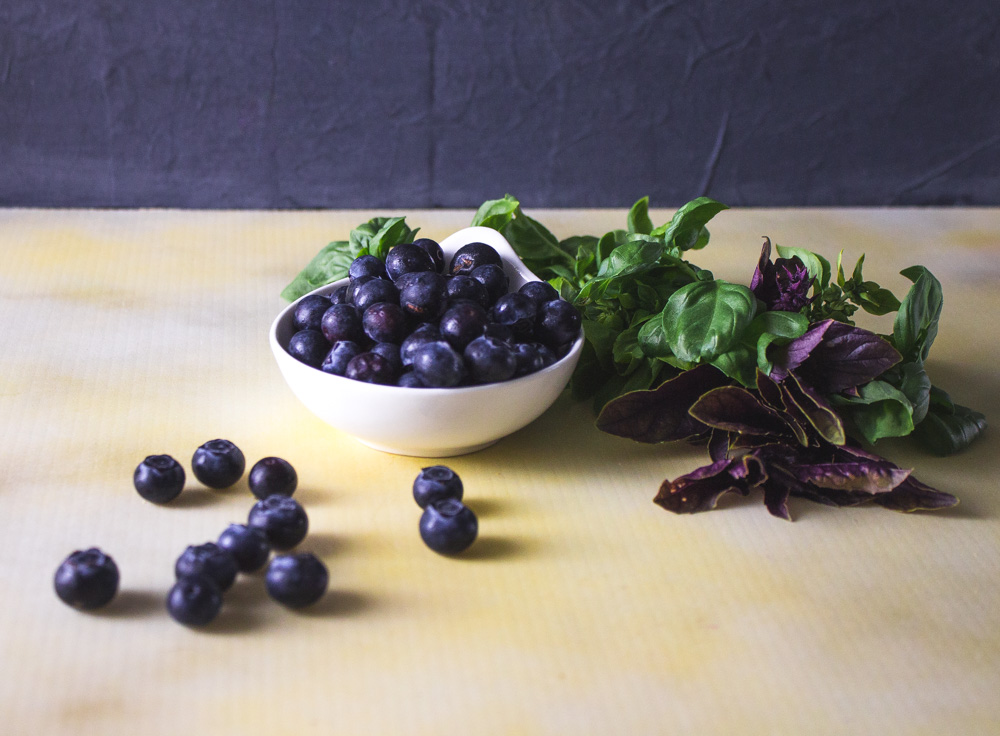 White bowl of blueberries and two types of basil on a yellow background