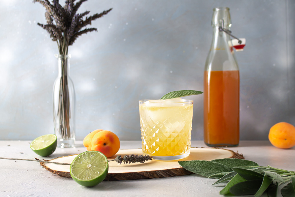 Apricot shrub gin spritz with bottle of shrub and fruit against a blue background