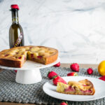 Slice of strawberry olive oil cake on a white plate with remainder of cake on stand with a bottle of olive oil.