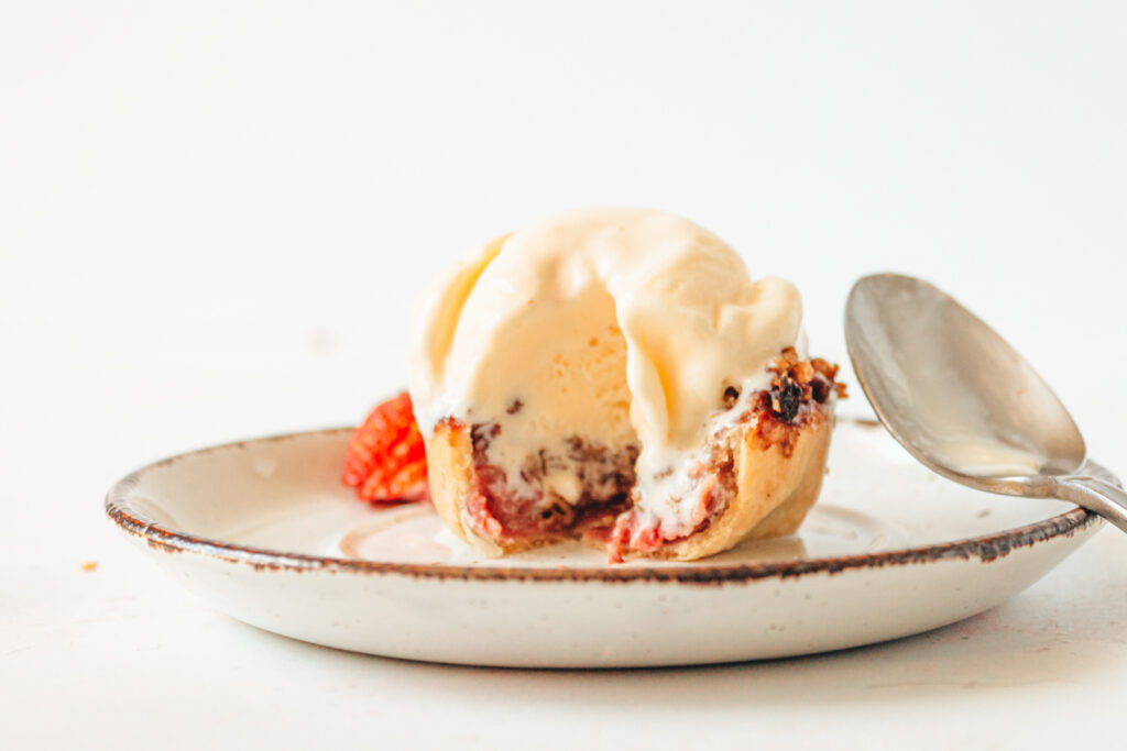 Handheld strawberry rhubarb crisp on a plate topped with ice cream with a bite taken out.