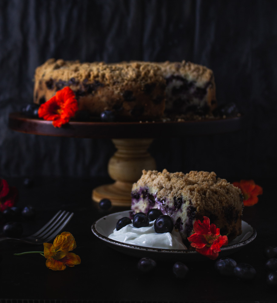 Slice on a plate with blueberry buckle on stand in background