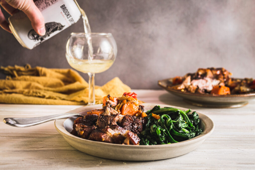 Tan plate with servings of braised pork and sweet potatoes and a side of sauteed greens. In the background a can of cider is being poured into a goblet and a serving dish with the rest of the braised meat.