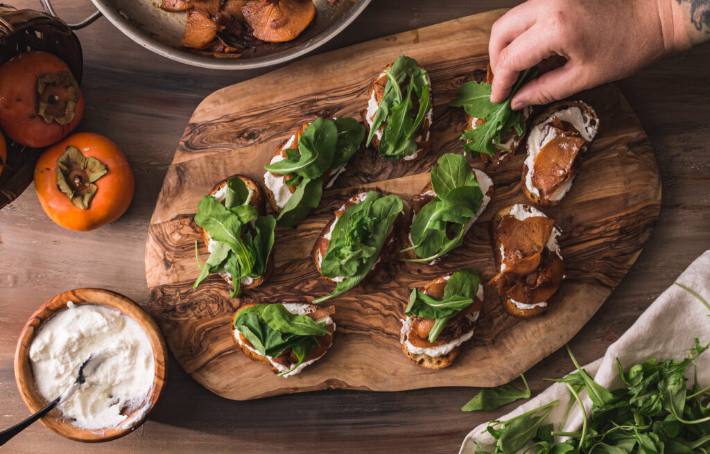 Overhead shot of white eprson's hand placing arugula on crostini. Other frinished crostini on the cutting board. Cutting board surrounded by fuyu persimmons, bowl of ricotta with spoon, and a plae napkin full of baby arugula.