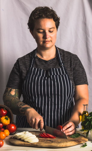 Non-binary person in a blue and white striped apron with short hair and tattoos cutting vegetables and cheese in front of a white background.
