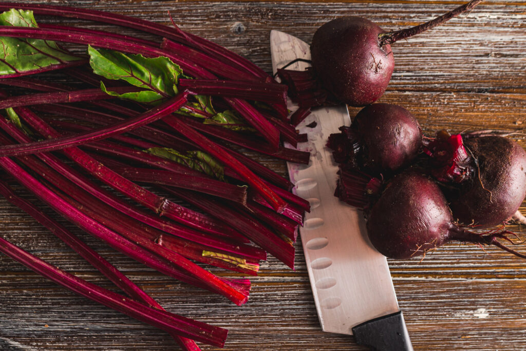 4 Red beets and stems on a wood board with a large knife. Beets and stems are cut just above the beet root.