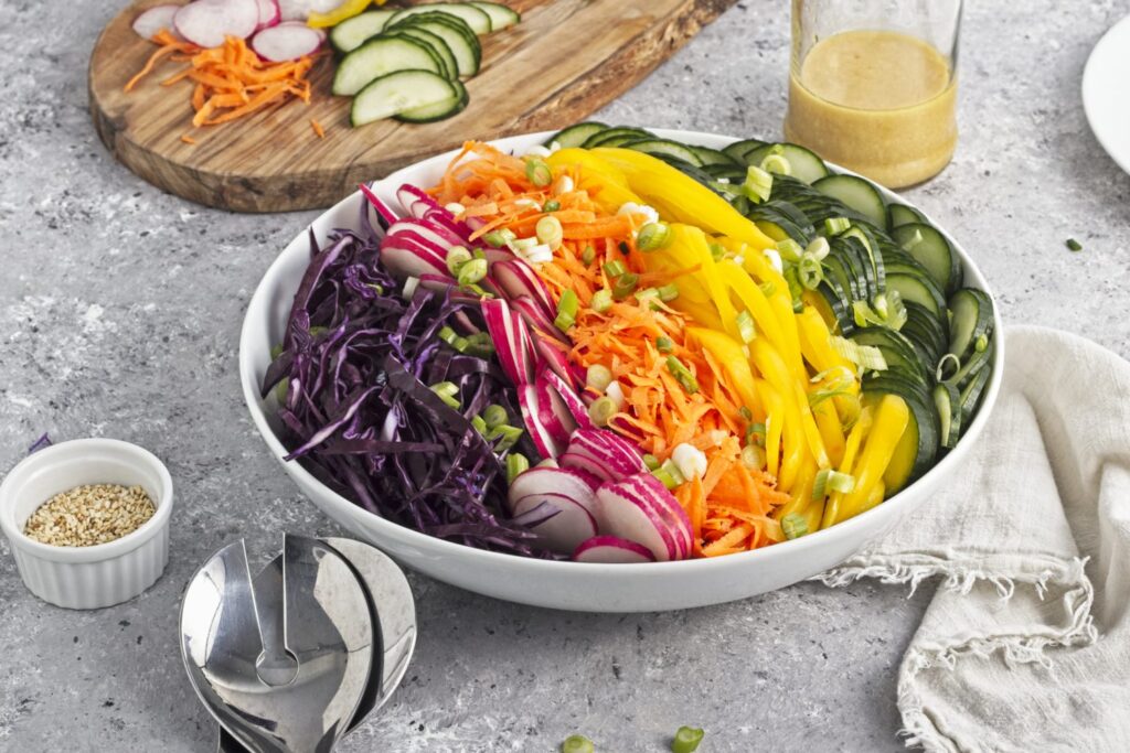 45 degree angled view of undressed rainbow coleslaw in a serving bowl with lines of purple cabbage, red radishes, orange carrots, yellow peppers, and green cucumber arranged in a rainbow. A jar of dressing and assort serving tools are arranged on the table scape.