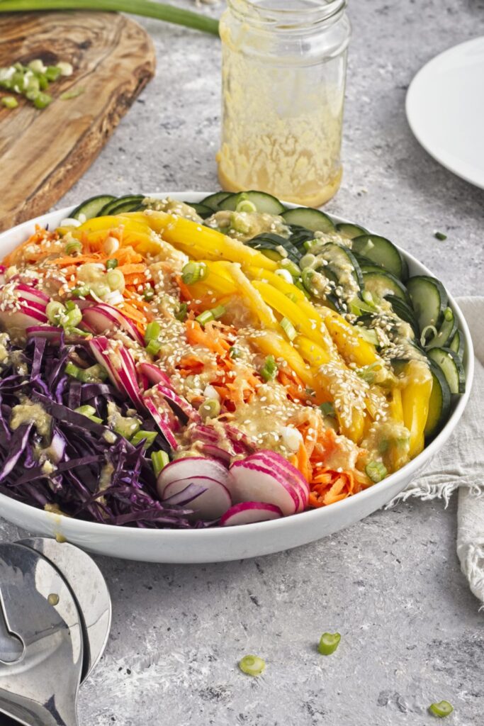 45 degree angled view of a serving bowl full of rainbow coleslaw on a tablescape with rows of purple cabbage, red radishes, carrots, yellow peppers, and cucumber.