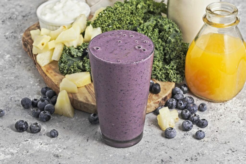 Slightly angled side view of Pint glass with a purple smoothie in front of a scatter of blueberries, pineapple chunks, kale, a dish of yogurt, a bottle of orange juice and a jar of protein powder