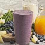 Pint glass with a purple smoothie in front of a scatter of blueberries, pineapple chunks, kale, a bottle of orange juice and a jar of protein powder
