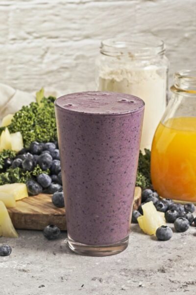 Pint glass with a purple smoothie in front of a scatter of blueberries, pineapple chunks, kale, a bottle of orange juice and a jar of protein powder