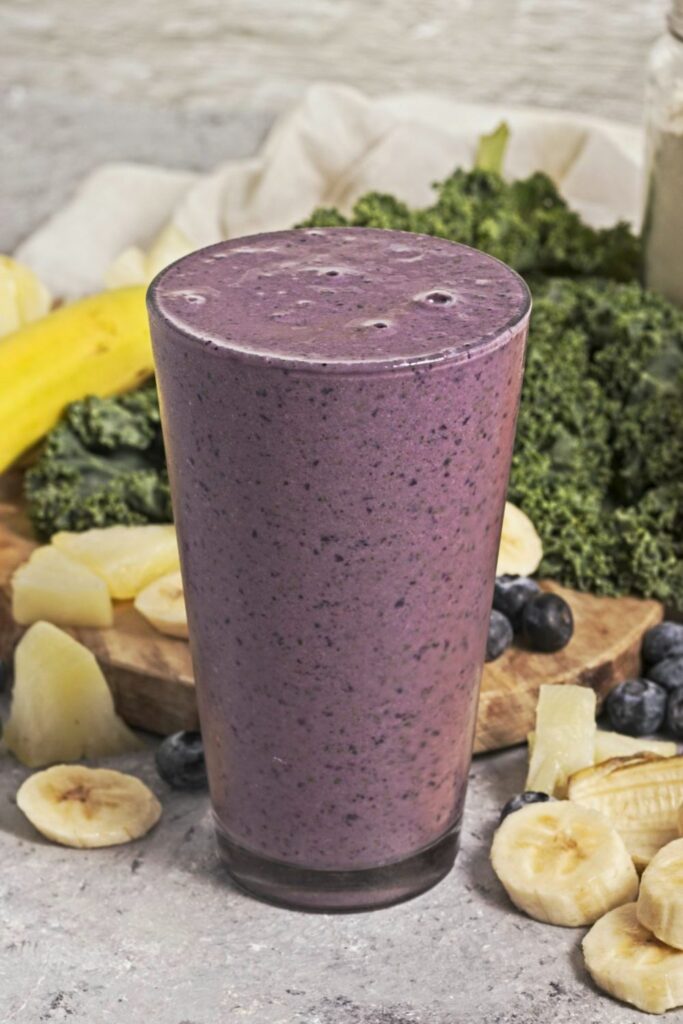 Pint glass with a purple smoothie in front of a scatter of blueberries, pineapple, sliced bananas, a bottle of orange juice and a jar of protein powder