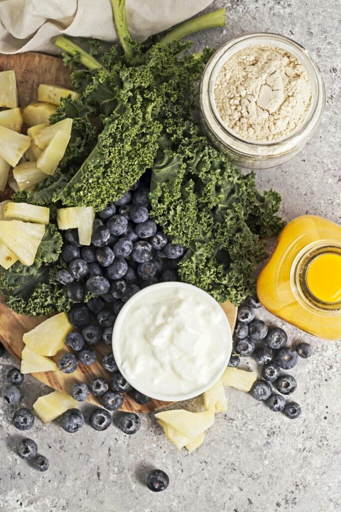 Overhead view of staged ingredients for the blue moon over brooklyn smoothie: blueberries, kale, pineapple chunks, a dish of yogurt, a bottle of orange juice and a jar of protein powder
