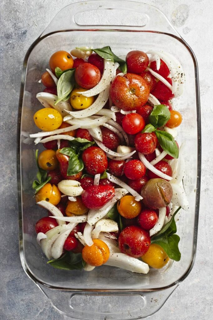 top down view of a baking dish filled with raw multicoloured cherry tomatoes, sliced onions, garlic cloves, and basil.