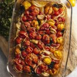 Top down view of a baking dish full of roasted multicoloured cherry tomatoes, onions, and basil. The baking dish is resting on a cutting board with chopped fresh basil to the side.