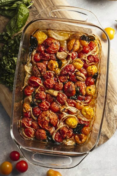 Top down view of a baking dish full of roasted multicoloured cherry tomatoes, onions, and basil. The baking dish is resting on a cutting board with chopped fresh basil to the side.