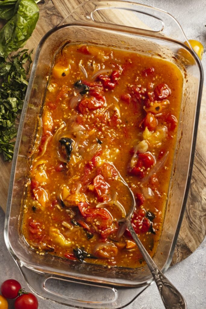 Top down view of a baking dish sitting on a a cutting board. The baking dish is full of tomato sauce all smashed together. Some fresh chopped basil is to the side of the baking dish.
