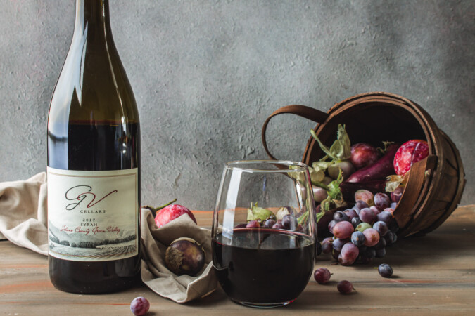 Several items on a wooden table. Bottle of GV Cellars syrah wine in infront of a tan napkin. A partially full glass of red wine. A basket on its side, spilling out various purple fruits and vegetables.