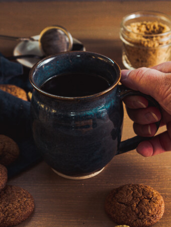 Hand holding a dark blue mug filled with dark liquid. On a wood table are molasses cookies, a tea strainer, and brown sugar in a jar.
