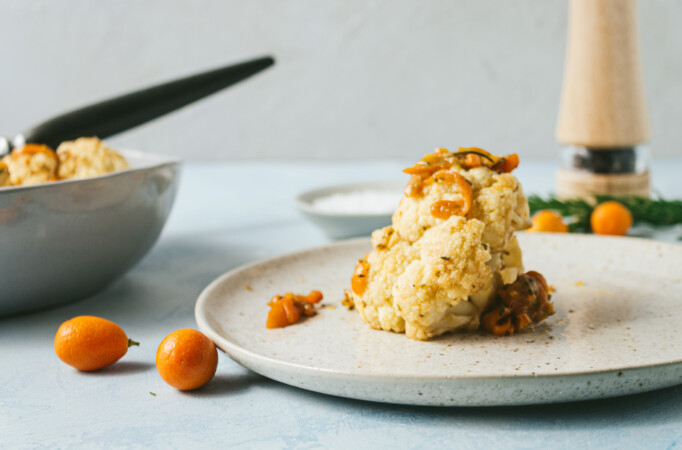 Cooked cauliflower with sliced kumquats on a tan plate. Bowl with serving spoon filled with more cauliflower in background. Scatter whole kumquats on blue surface surounding it.