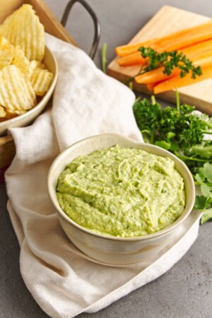 Bowl of light green dip with a second larger bowk of potato chips behand it and a cutting board with carrot sticks to the side. Scattered parsley and cilantro are between the dishes and a off-white napking is drapped decoratively.